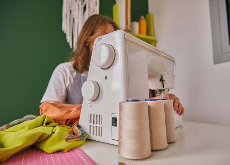 A person sewing at a white sewing machine with thread spools, bright fabric, and cutting mat on a tidy craft workspace.
