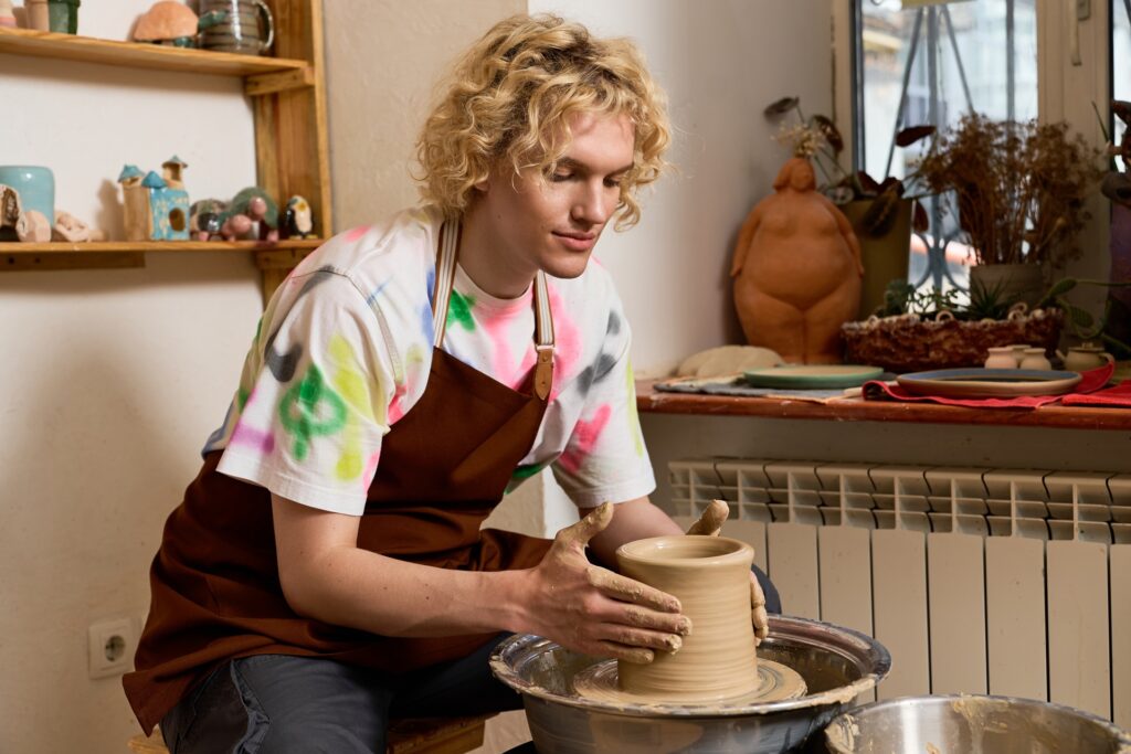 A young man working on a pottery wheel. He has a pot in the middle of being made spinning in his hands.