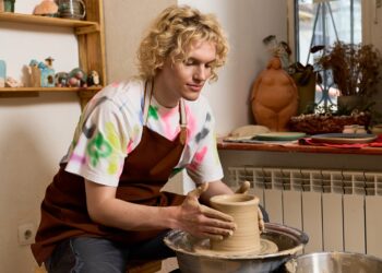 A young man working on a pottery wheel. He has a pot in the middle of being made spinning in his hands.