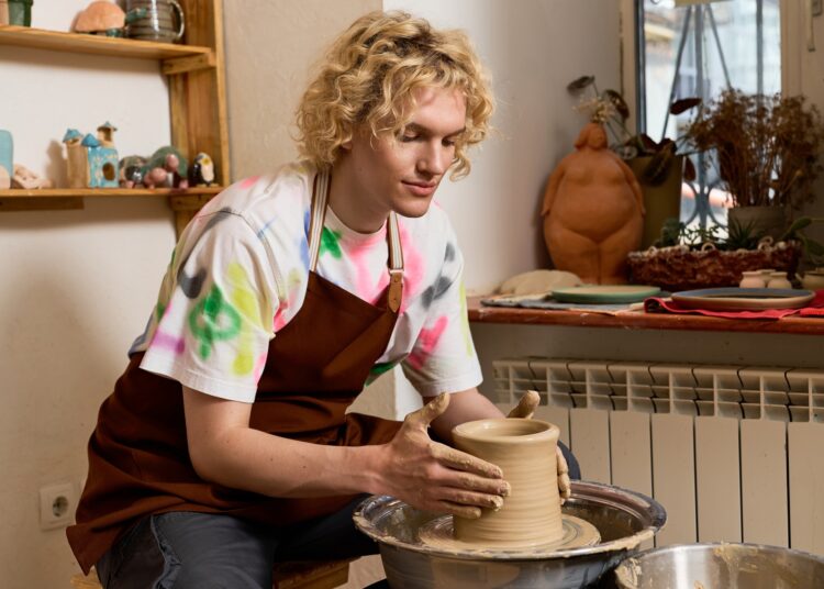 A young man working on a pottery wheel. He has a pot in the middle of being made spinning in his hands.