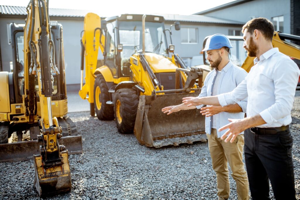A contractor in a hard hat and a businessman standing near a row of heavy machinery. Both are pointing at one machine.