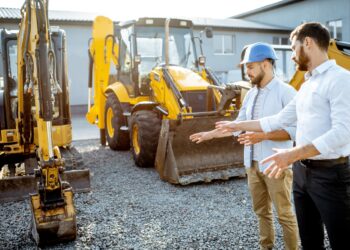 A contractor in a hard hat and a businessman standing near a row of heavy machinery. Both are pointing at one machine.