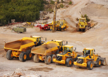 An overhead view of multiple yellow tractors and dump trucks parked in a row at a construction site.