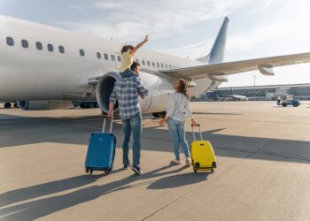 Two adults and a child walking toward a large airplane on an airport tarmac while pulling blue and yellow suitcases.