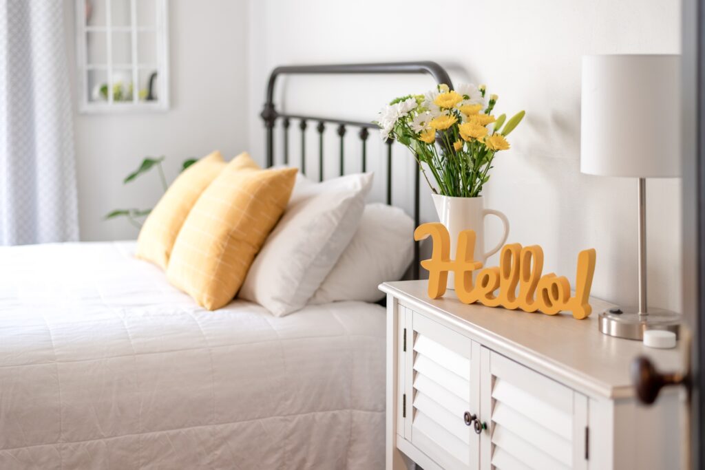 A bright guest bedroom with white bedding, yellow pillows, flowers in a vase, a lamp, and a cheerful hello sign on a cabinet.