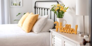 A bright guest bedroom with white bedding, yellow pillows, flowers in a vase, a lamp, and a cheerful hello sign on a cabinet.