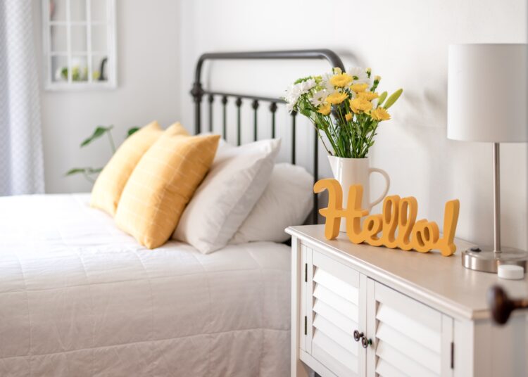 A bright guest bedroom with white bedding, yellow pillows, flowers in a vase, a lamp, and a cheerful hello sign on a cabinet.