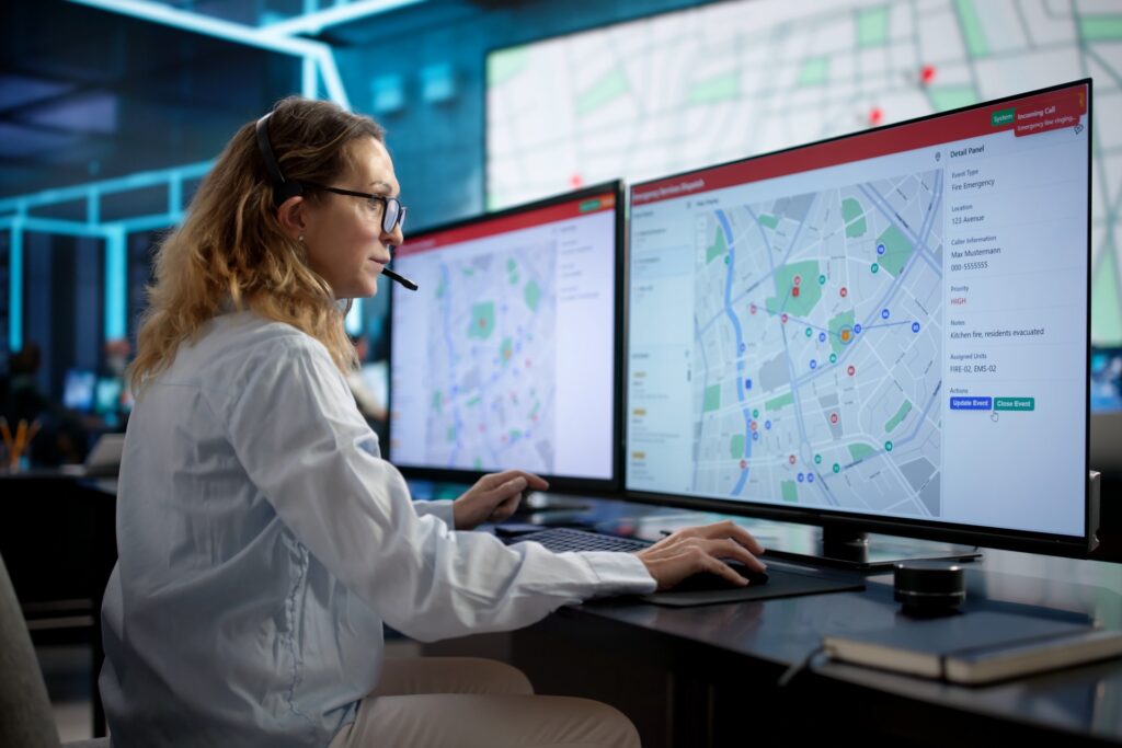 A woman wearing a headset sits at a desk in front of two very large monitors. Two maps cover both screens.
