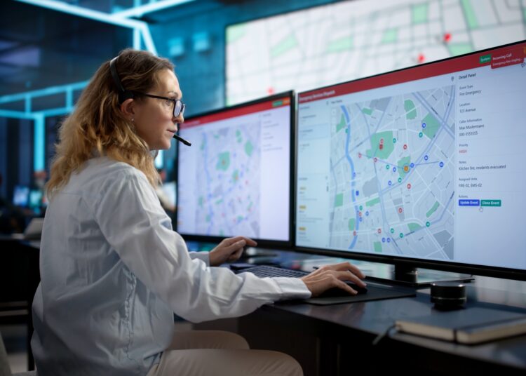 A woman wearing a headset sits at a desk in front of two very large monitors. Two maps cover both screens.
