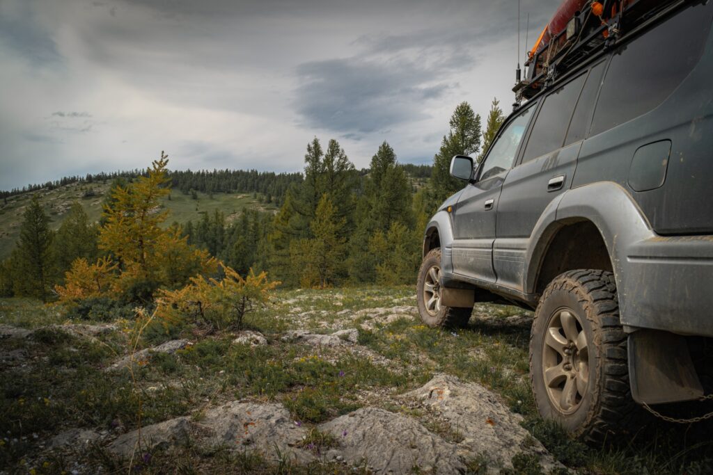 An overland SUV with rugged tires is parked on rocky terrain overlooking a forested mountain landscape under cloudy skies.