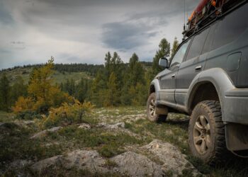 An overland SUV with rugged tires is parked on rocky terrain overlooking a forested mountain landscape under cloudy skies.